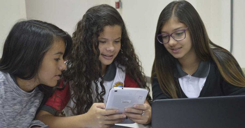 Tres niñas miran una tablet en un salón de clases.