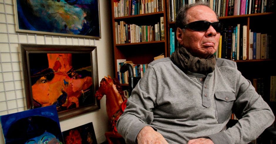 Rafa Fernández con unos lentes oscuros y camisa gris, sentado en su casa, en su biblioteca, con tres obras suyas de fondo