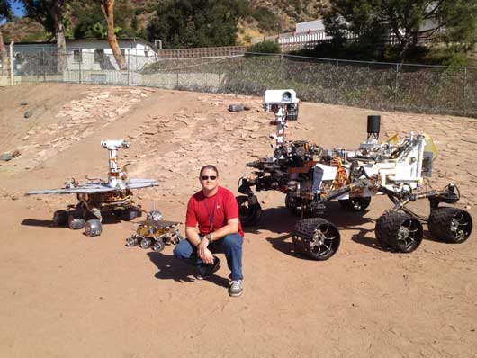 Alex Mora, técnico aeroespacial del Laboratorio de Propulsión a Chorro de la NASA, posa con varios Mars Rover.