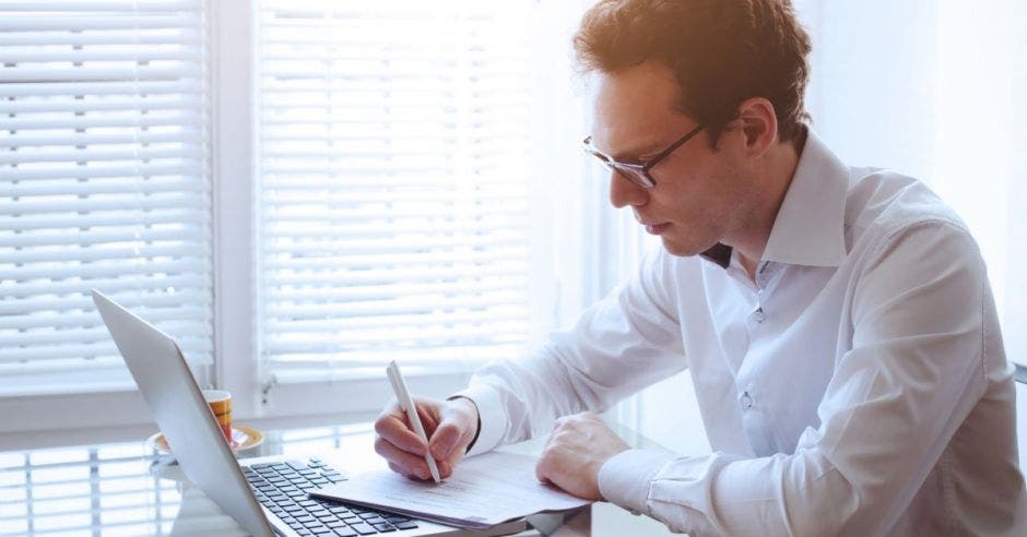 Un hombre revisando el computador mientras escribe en algunos papeles que tiene en su escritorio