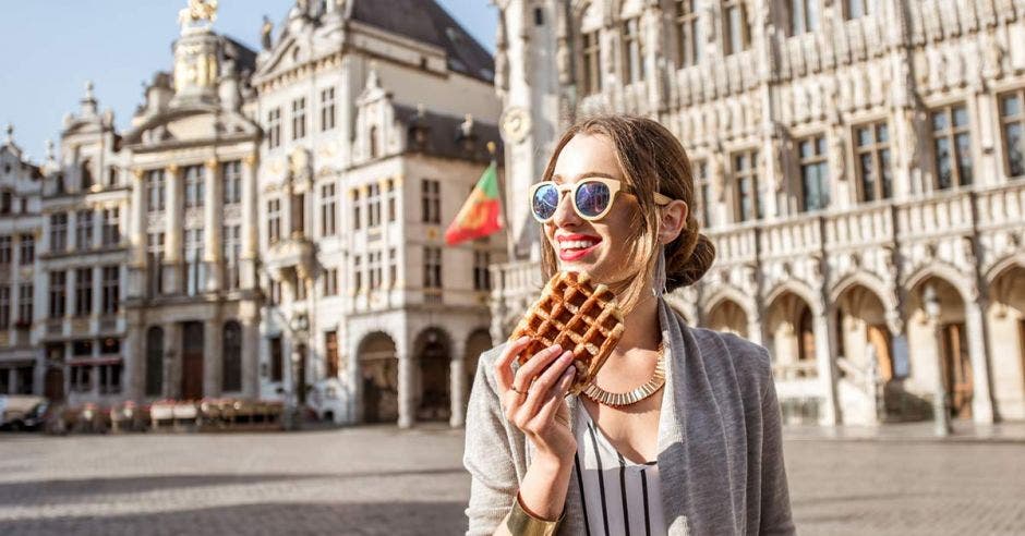 Una mujer sonriendo con un Waffle en la mano, en el fondo unos edificios en Bélgica