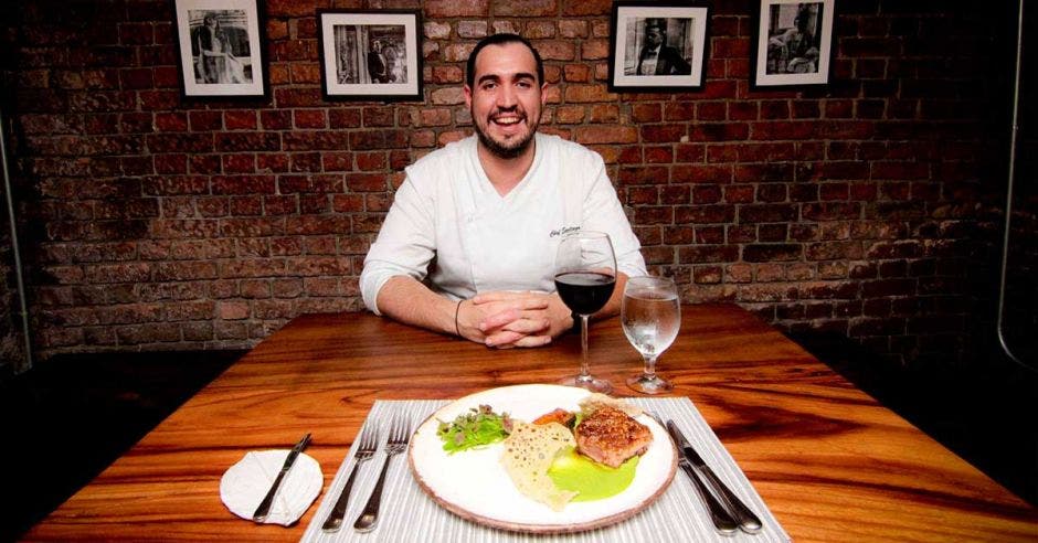 El chef, hombre de pelo negro, con camisa blanca, sentado frente a uno de sus platillos y una copa de vino