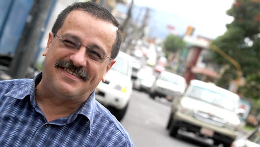 Hombre con anteojos, bigote, camisa cuadros en la calle carros detrás