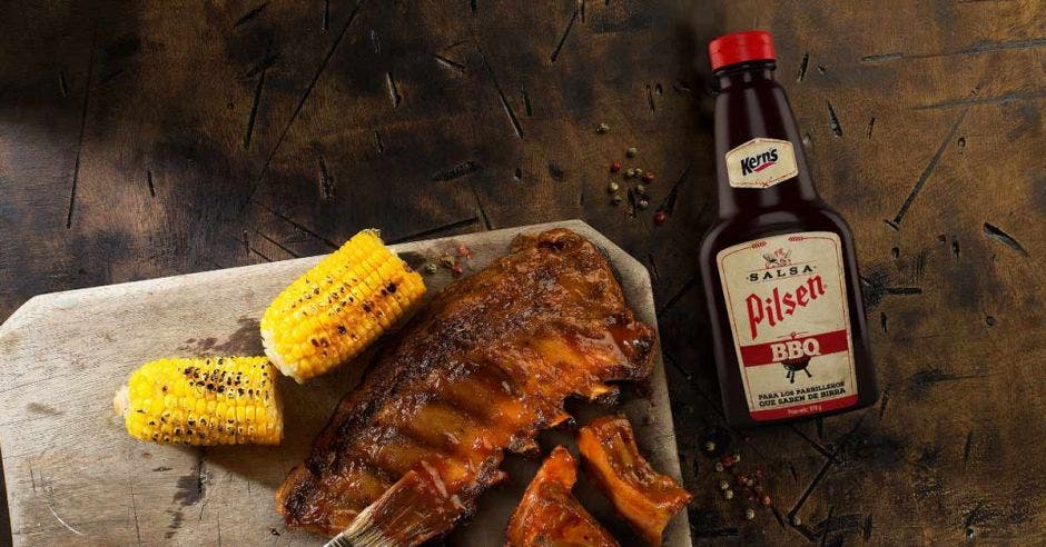 botella de Pilsen BBQ junto a un trozo de carne y dos de maíz