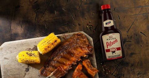 botella de Pilsen BBQ junto a un trozo de carne y dos de maíz