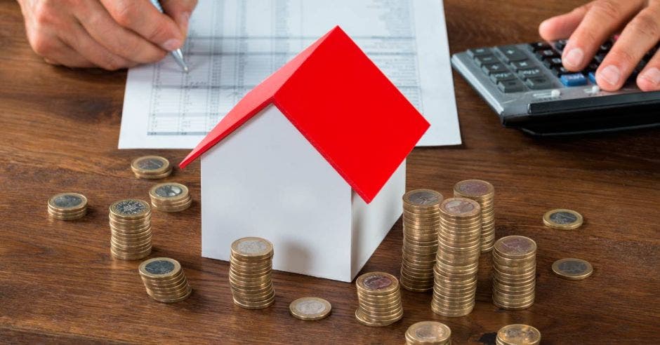 Foto de una casa pequeña sobre una mesa y a su alrededor monedas apiladas