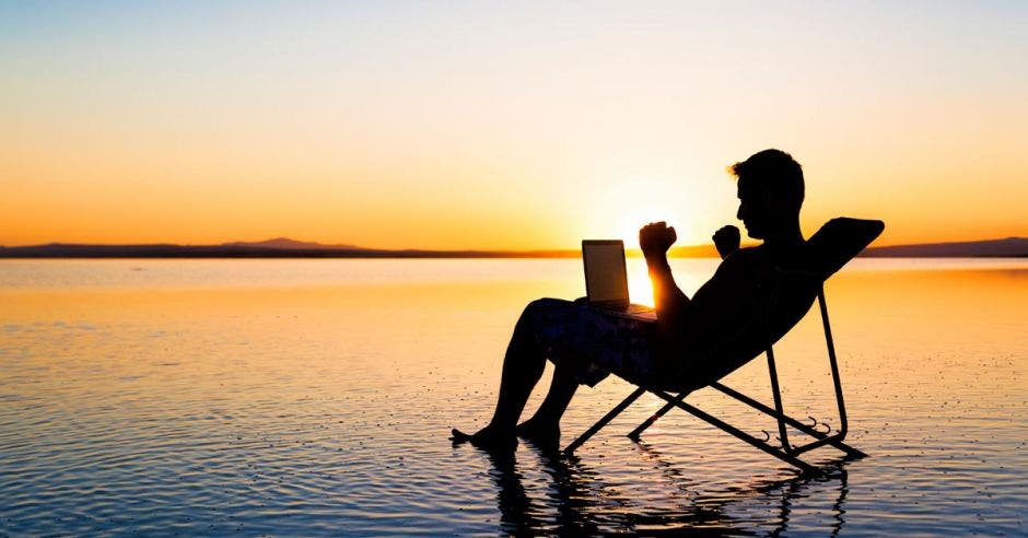 Persona en silla de playa, sentada frente al mar, en un atardecer, con computadora en sus piernas y brazos asemejando celebración