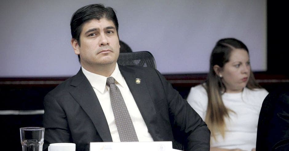 El presidente Carlos Alvarado sentado en una mesa dando una conferencia, mientras que un panelista se encuentra en la parte trasera.