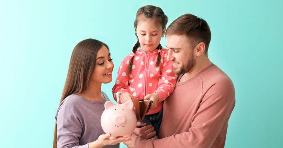 familia de tres personas, una mujer, y un hombre alzando a una niña, sostienen una alcancía de chancho rosada