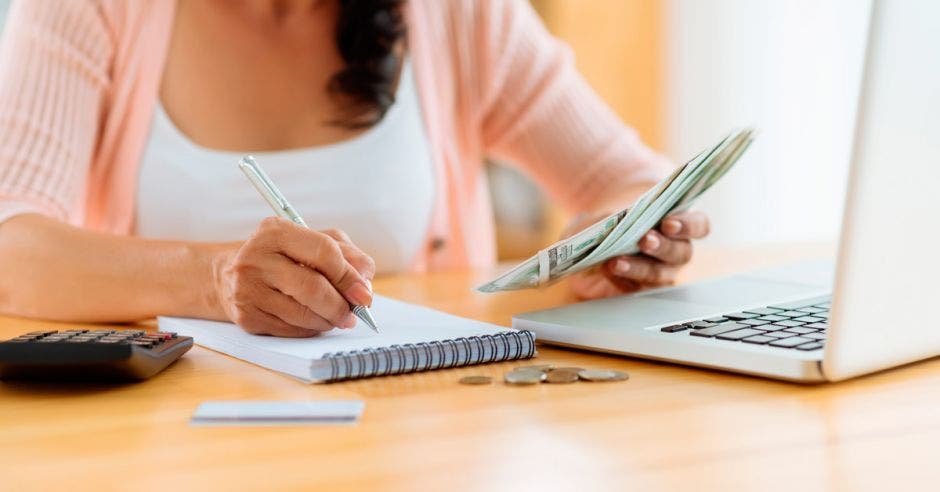 mujer realiza cálculos de su presupuesto con su computadora, libreta y calculadora. En la mano derecha sostiene dinero.