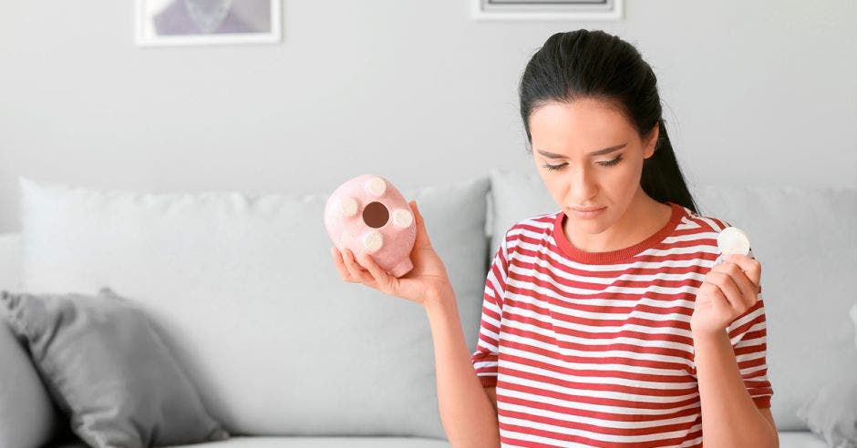 mujer realiza cálculos con la calculadora y libreta con una alcancía de chancho vacío en una mano y en la otra, una moneda