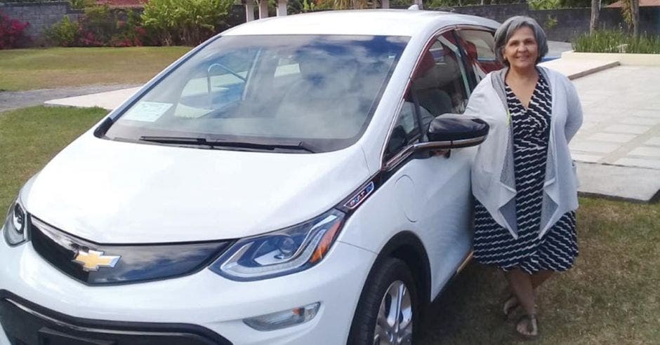 Una mujer de avanzada edad posa junto a un carro marca chevrolet color blanco