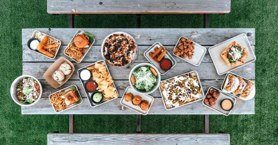 Una mesa llena de platillos de comida variados, entre ellos hamburguesas, papas fritas, emparedados y demás comidas artesanales