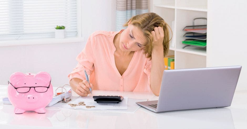 mujer pensativa haciendo un presupuesto frente a la computadora y una calculadora, y una alcancía de cerdito rosada al lado