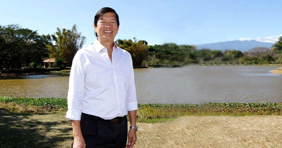 Un adulto asiático viste camisa blanca y pantalones, con el lago de la sabana de fondo