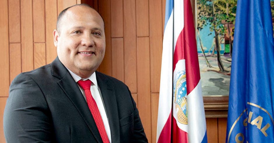 Un hombre posa junto a dos banderas, una de costa rica y otra color azul con el logo de la CCSS