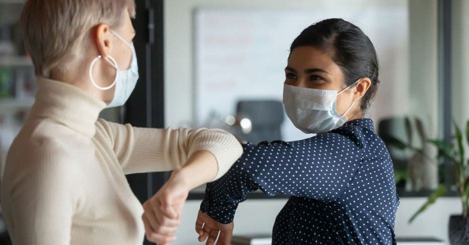 Vemos a dos mujeres con mascarillas dándose codo con codo