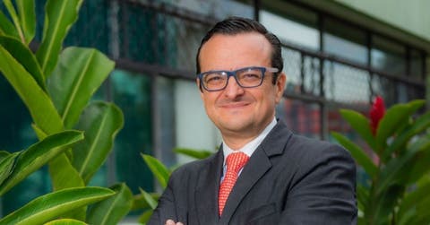 Un hombre con anteojos sonríe frente a un edificio color gris