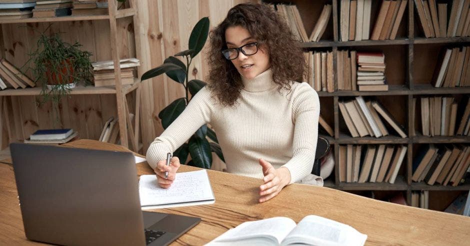 Muchacha de lentes tomando clase virtual en su laptop con libros abierto a un lado