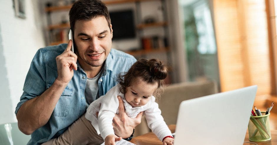 Un padre de familia sosteniendo a una bebé mientras hace teletrabajo