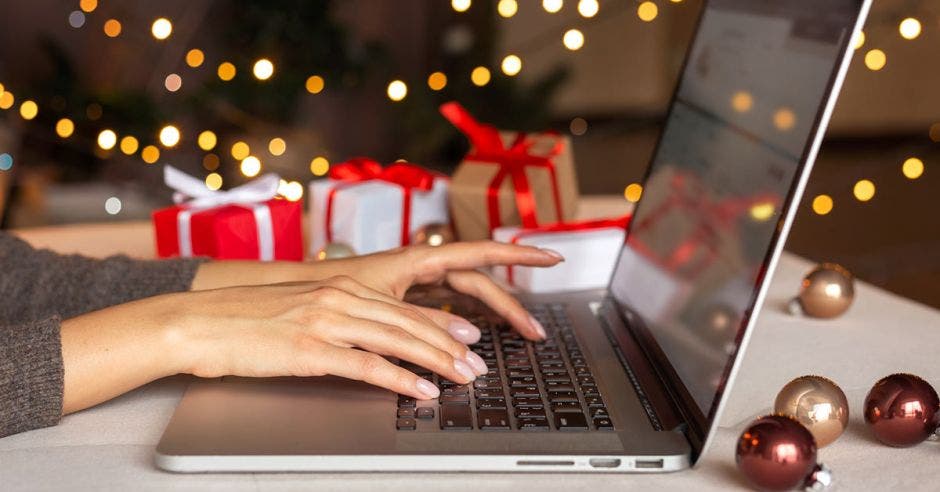 manos de mujer presionando teclas de una laptop con un árbol de navidad y regalos de fondo