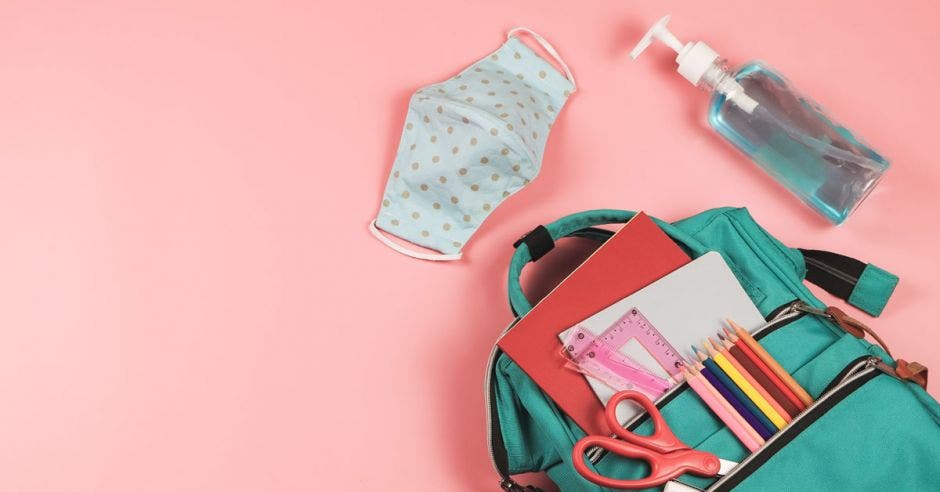 mochila con artículos escolares sobre fondo rosa, al lado de un cubrebocas y gel antibacterial