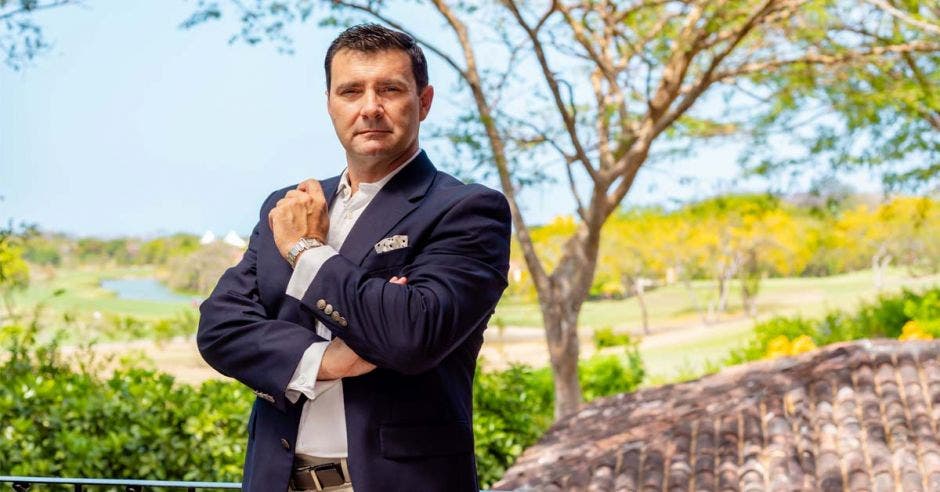 un hombre de saco azul posa sobre un fondo verde con naturaleza y flores