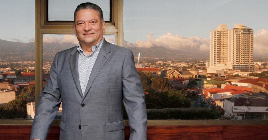 un hombre con saco gris frente a una ventana con una ciudad de fondo