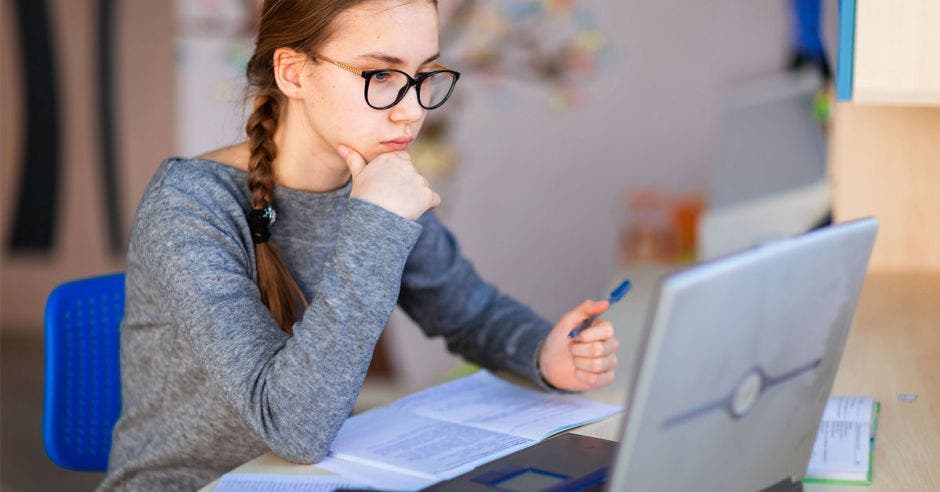 mujer joven con lentes y sueter color gris, sosteniendo un bolígrafo azul en la mano derecha frente a una laptop