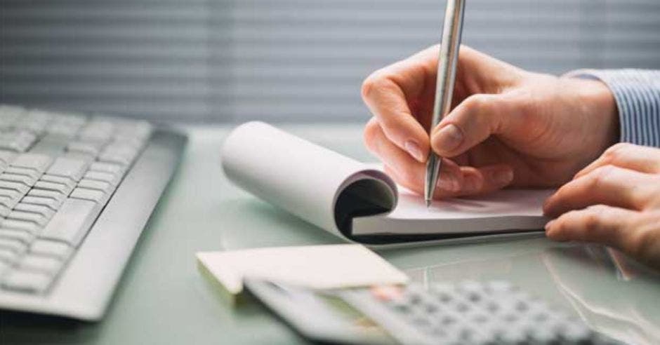 mano sosteniendo un bolígrafo anotando en una libreta junto a un computador