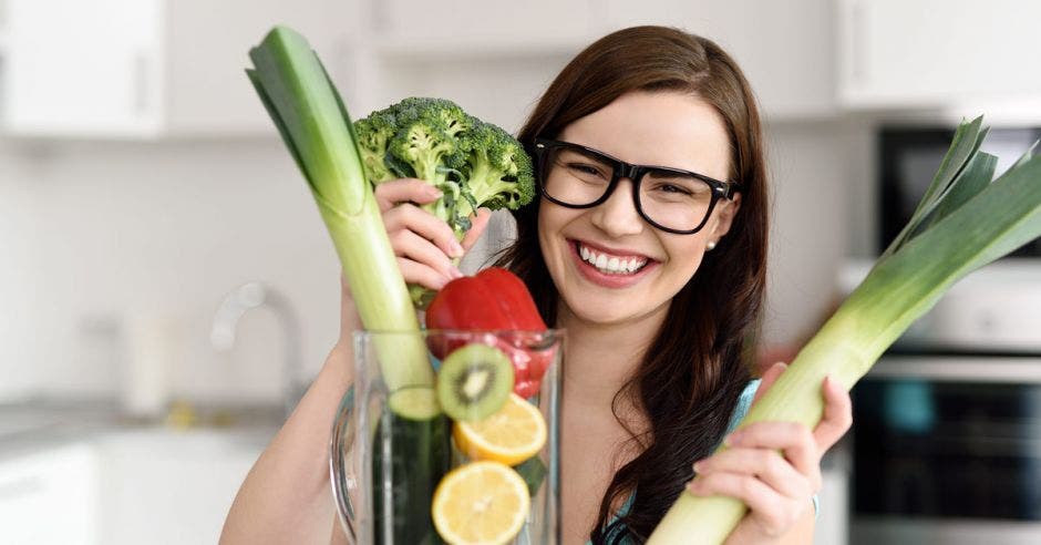 mujer joven con lentes, sonriendo, sosteniendo vegetales frente a una licuadora