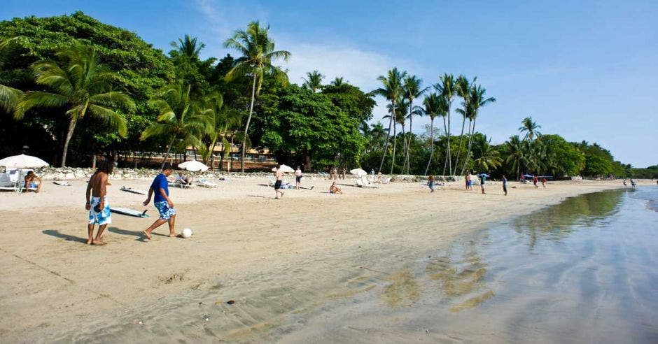 dos personas juegan al fútbol en una playa. Varias personas alrededor.