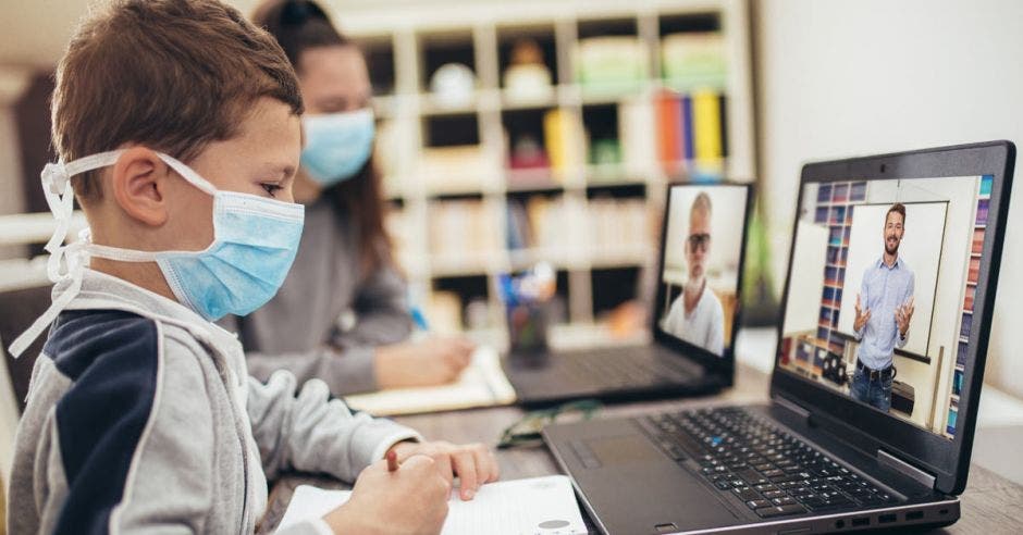 Unos niños con mascarilla viendo en una computadora al docente que teletrabaja