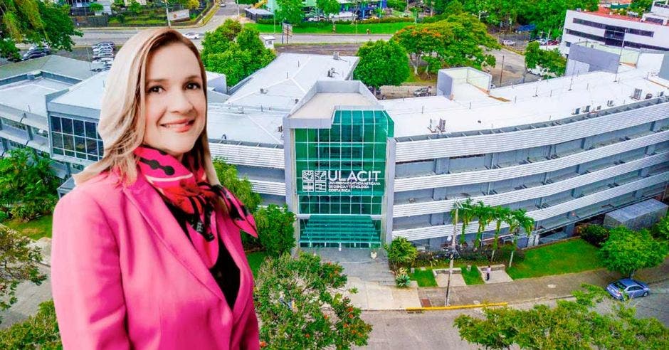 mujer rubia con saco rosa y mascada rosa, de fondo instalaciones de una universidad