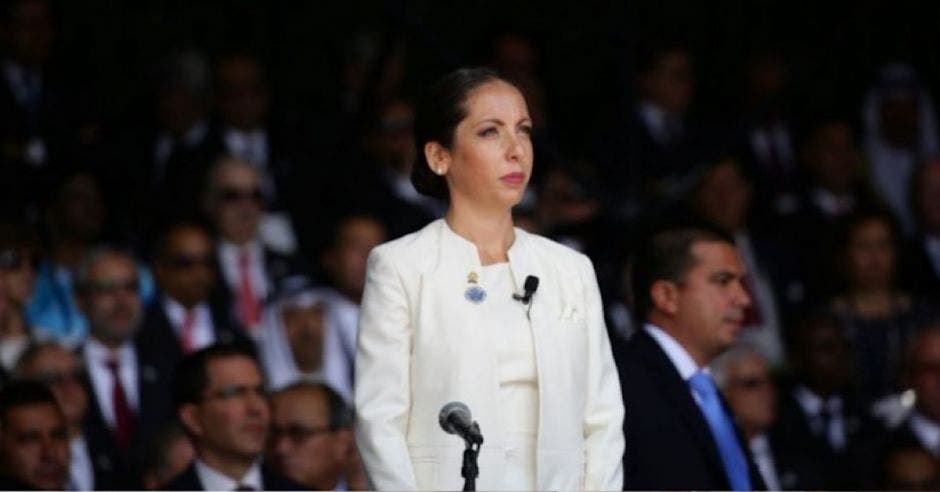 mujer joven con saco y blusa blanca de pie frente a un micrófono