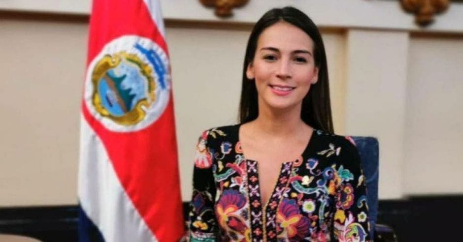 mujer joven de cabello liso negro con blusa negra floreada, detrás bandera nacional