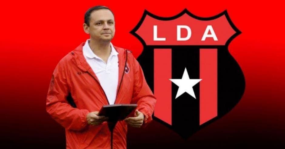 hombre con chamarra roja y playera blanca detrás logotipo de Alajuelense