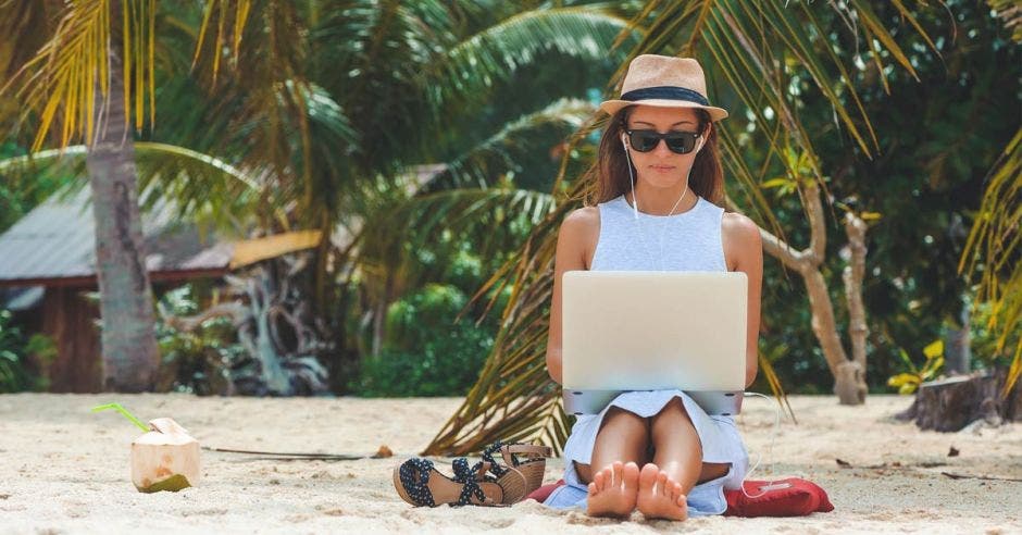 Joven mujer en vestido, gafas de sol y sombrero, trabajando en laptop en la playa, en el fondo de las palmeras