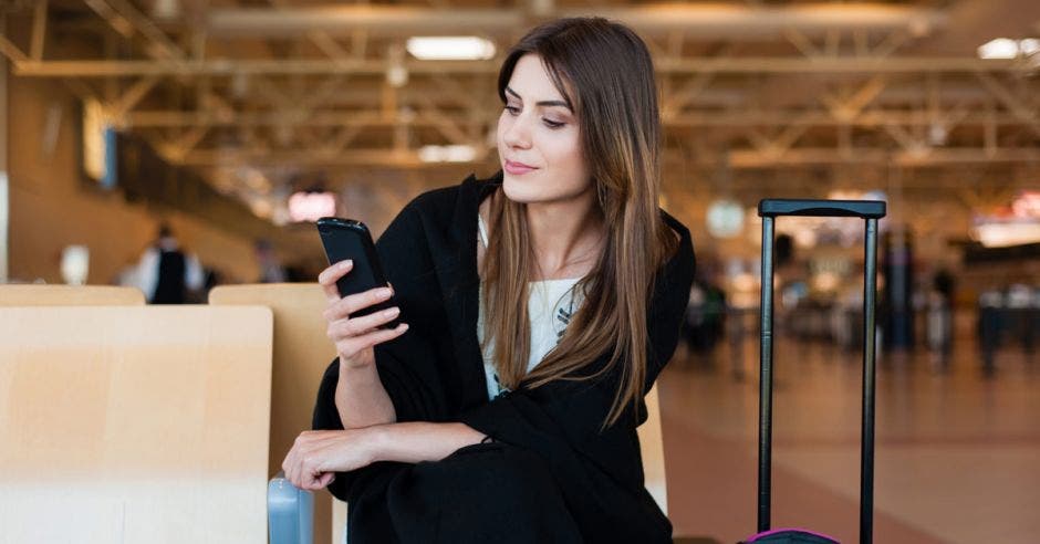 Concepto de viaje aéreo con una joven mujer informal sentada con maleta de equipaje de mano y revisando su celular