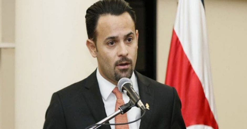 un hombre habla frente a un micrófono, con la bandera de Costa Rica de fondo