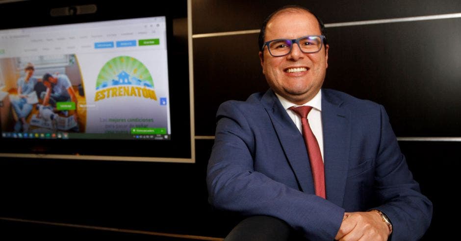 hombre con lentes, traje azul y corbata roja con brazo recargado sobre sillón