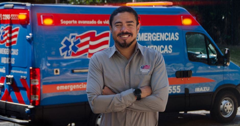 hombre de cabello negro y barba, con camisa gris y brazos cruzados, detrás una ambulancia