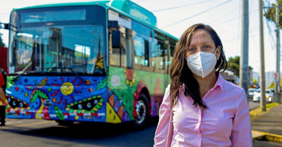 una mujer con mascarilla blanca y blusa rosada posa en frente de un autobús colorido