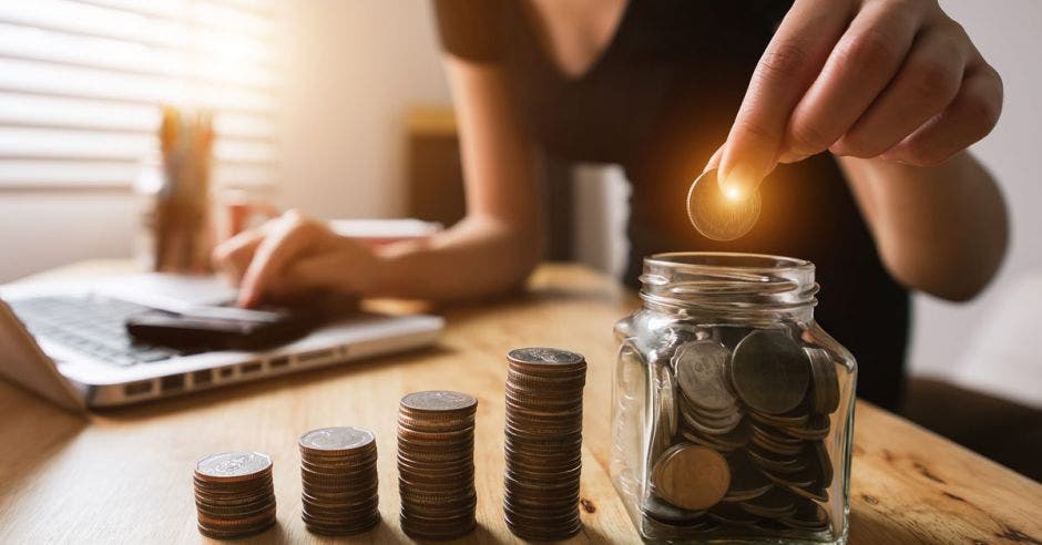 mujer depositando moneda en tarro de vidrio junto a torres de monedas de diferente tamaño