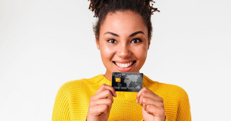 mujer sonriendo, con sueter amarillo sosteniendo una tarjeta de débito con las dos manos