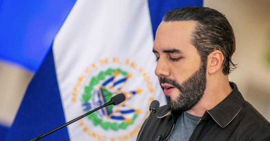 un hombre de barba habla frente a un micrófono. De fondo la bandera de El Salvador.