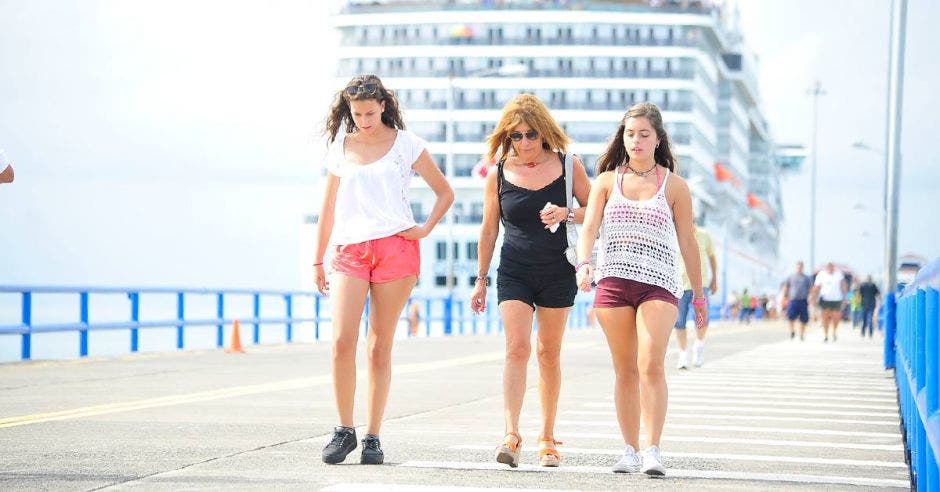 tres mujeres caminan por un puerto. Un crucero encallado en el fondo.