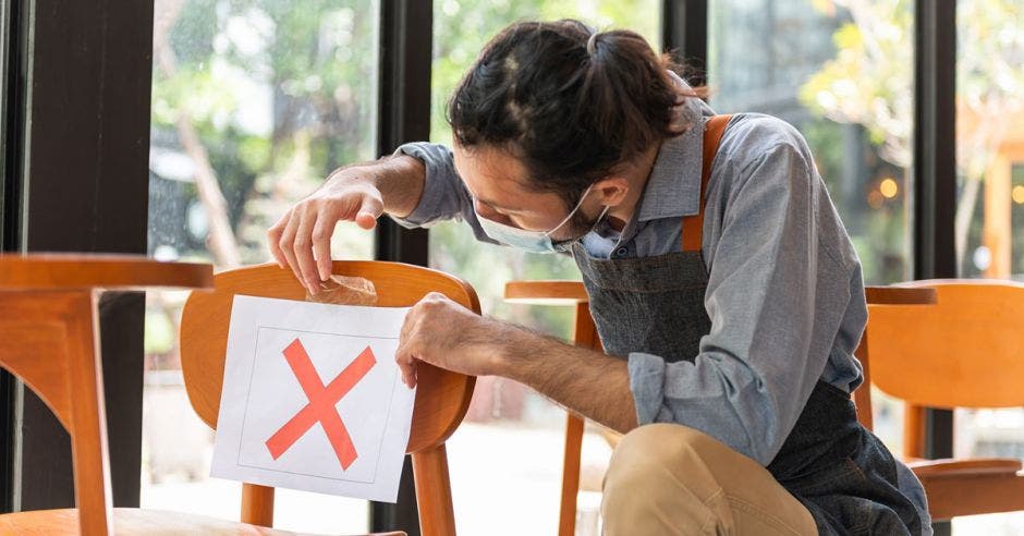 Un camarero asiático pone una señal cruzada en la silla del restaurante o cafetería para hacer distanciamiento social para evitar la propagación del coronavirus