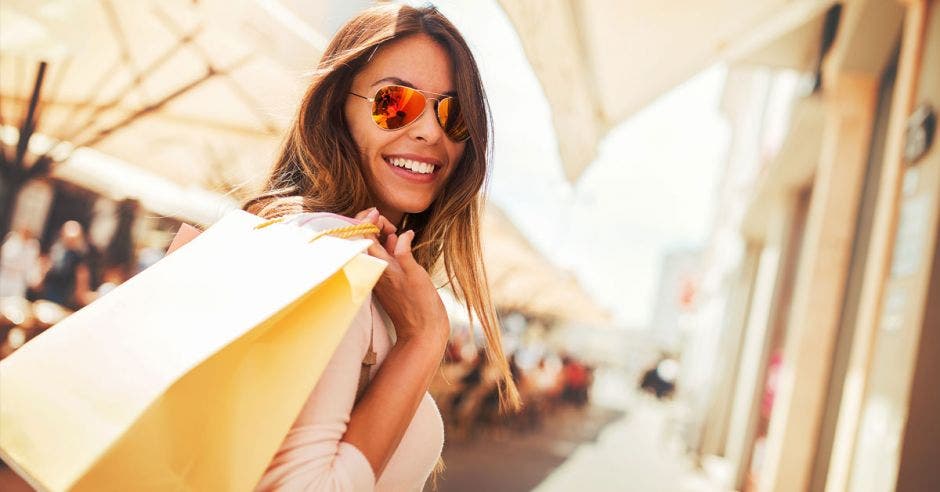 mujer de cabello castaño con lentes de sol, sonriendo y sosteniendo bolsas con la mano sobre el hombro