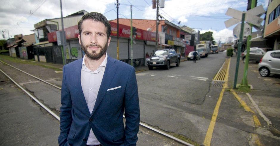 un hombre blanco de barba con camisa blanca y saco azul de pie frente a la línea del tren en una calle de asfalto.